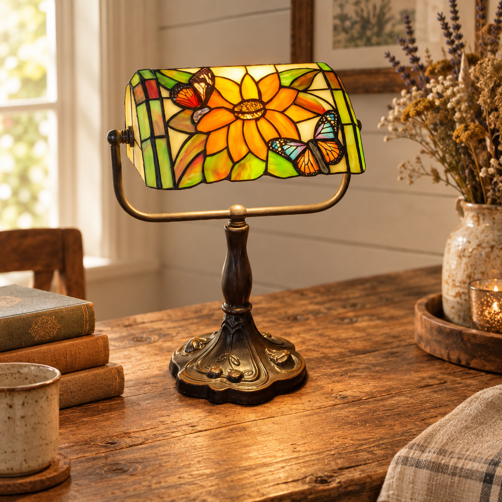 A Tiffany-style stained glass table lamp with a floral and butterfly design, mounted on an ornate dark metal base and shown turned on on a wooden table.