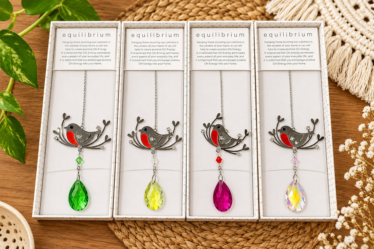 Four boxed bird light catcher displayed side by side on a woven mat. Each light catcher has a silver bird charm with a small red accent and a dangling teardrop crystal in a different colour: green, yellow, pink, and clear.