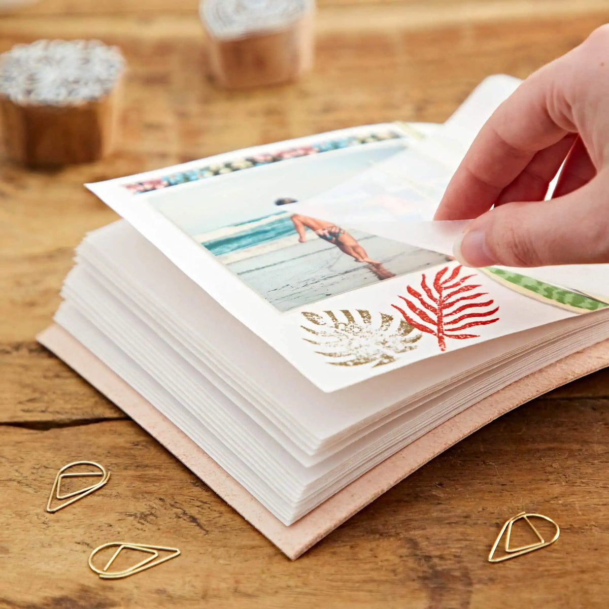 A hand is peeling back a printed photo sheet from a stack of white sheets placed on a tan leather journal or book, with gold paperclips on a wooden tabletop.