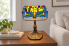 A Tiffany-style stained glass desk lamp featuring a dragonfly design, displayed on a dark wood side table in a home setting.