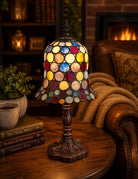 A stained glass table lamp with a colorful circular pattern and an ornate dark bronze base, styled on a wooden table next to a leather armchair in a cozy living room setting.
