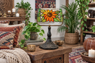 A decorative table lamp with a stained-glass style sunflower shade and a dark metal base, styled on a wooden side table in a cozy, rustic living room setting with plants, pillows, and candles.