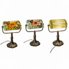 Three views of a Tiffany-style stained glass desk lamp with a floral pattern, featuring an ornate dark metal base and a yellow-green striped shade, shown lit on a wooden table.