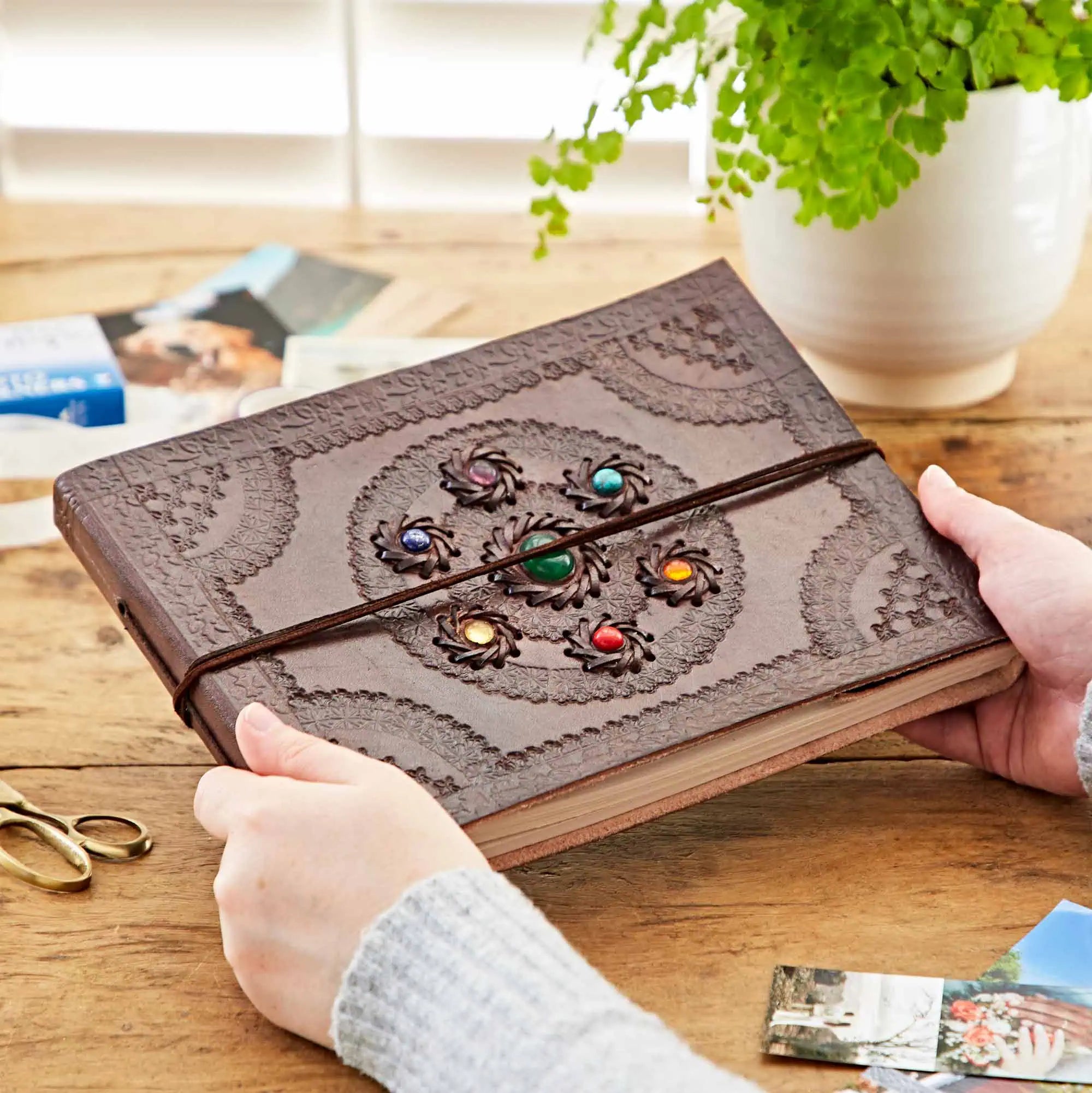 A person holds a brown embossed leather journal with a circular floral pattern and colorful gemstone accents on the cover, tied with a dark elastic band, on a wooden table with a potted plant in the background.