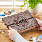 A person holds a brown embossed leather journal with a circular floral pattern and colorful gemstone accents on the cover, tied with a dark elastic band, on a wooden table with a potted plant in the background.