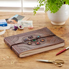A brown embossed leather journal with a decorative center medallion and colorful rhinestone accents, tied with a black elastic band, displayed on a wooden desk with stationery and a potted plant.