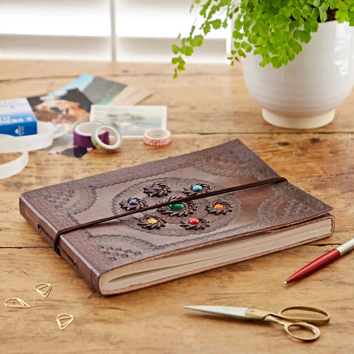 A brown embossed leather journal with a decorative center medallion and colorful rhinestone accents, tied with a black elastic band, displayed on a wooden desk with stationery and a potted plant.