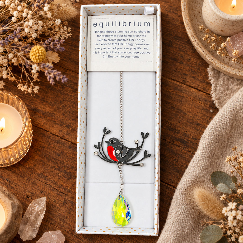 A bird-shaped hanging sun catcher displayed inside an open white gift box on a wooden table, surrounded by dried flowers, crystals, and lit candles. The sun catcher has a black bird body with a red chest, a silver chain, and a yellow-green faceted glass pendant.