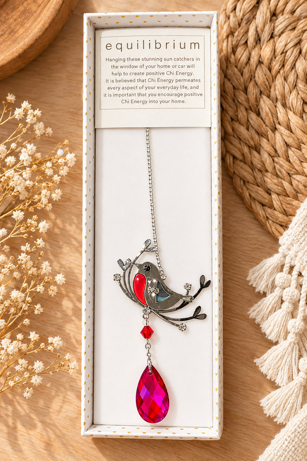 A boxed necklace with a silver-toned branch and bird pendant, featuring a bright red gemstone and a large pink teardrop crystal, displayed in a white gift box labeled 'equilibrium' on a warm neutral background with dried flowers and woven decor.