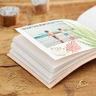 A stack of printed paper cards or postcards on a wooden table, with one card showing a beach photo and decorative palm motifs. Gold paperclips are placed nearby, and blurred containers appear in the background.