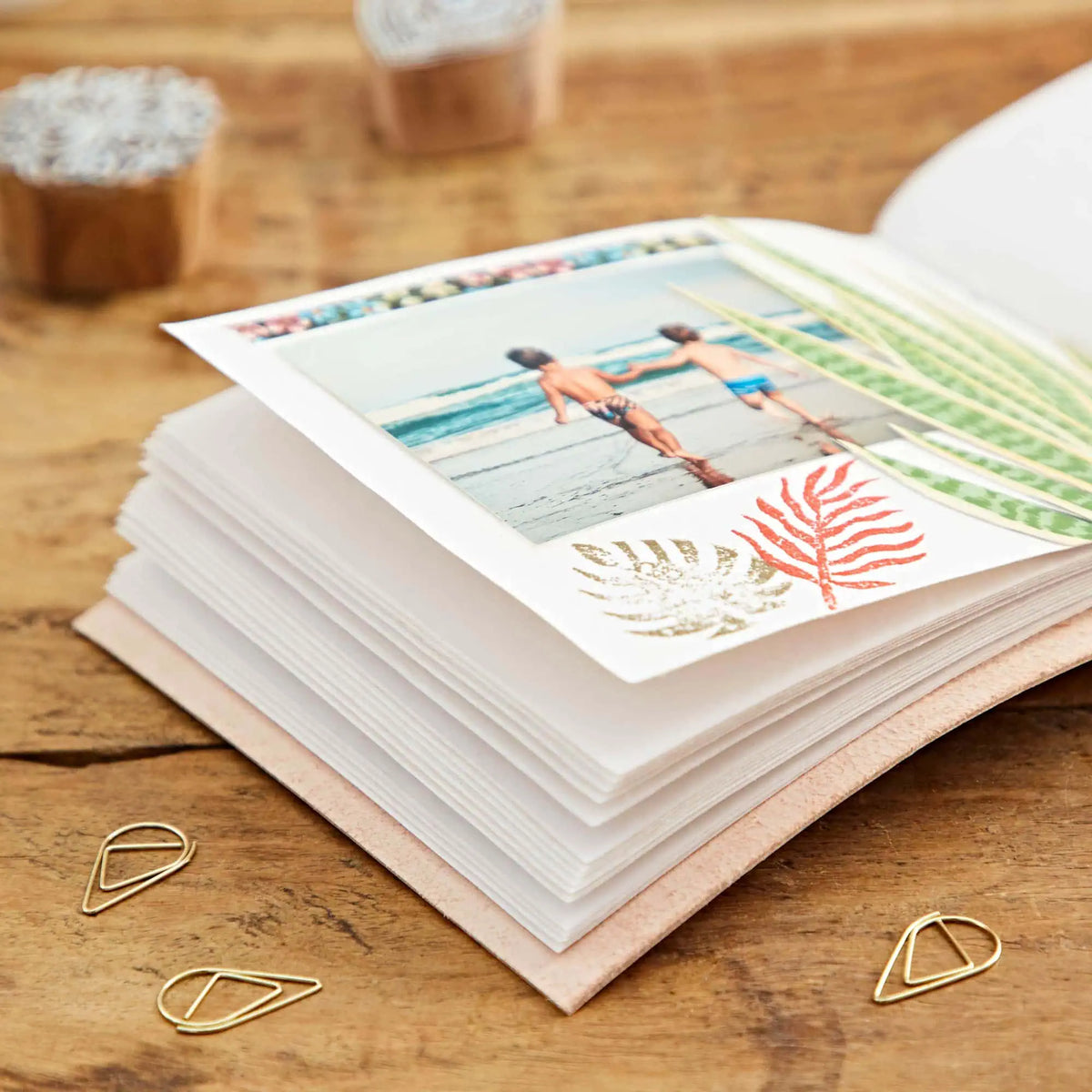 A stack of printed paper cards or postcards on a wooden table, with one card showing a beach photo and decorative palm motifs. Gold paperclips are placed nearby, and blurred containers appear in the background.