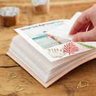 A hand is peeling back a printed photo sheet from a stack of white sheets placed on a tan leather journal or book, with gold paperclips on a wooden tabletop.