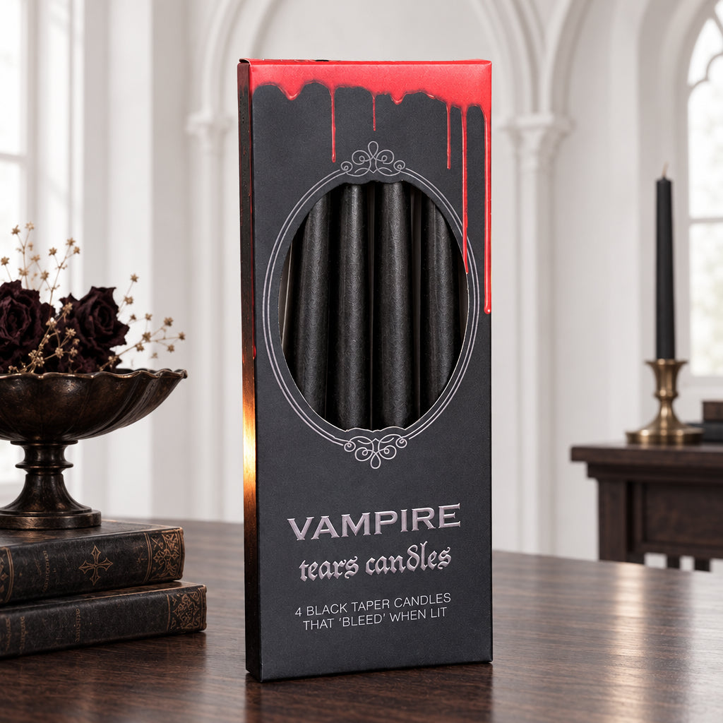 A boxed pack of vampire taper candles stands on a dark wooden surface, flanked by two lit black candles against a black fabric backdrop. The package is dark gray with a circular window showing black candles inside and the text 'VAMPIRE taper candles' and '4 BLACK TAPER CANDLES THAT BLEED WHEN LIT'.