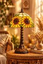A Tiffany-style stained glass table lamp featuring two large sunflower faces on a dome-shaped shade, with green leaves and a dark metal tree-like base, shown on a wooden table in front of a dark leather chair.